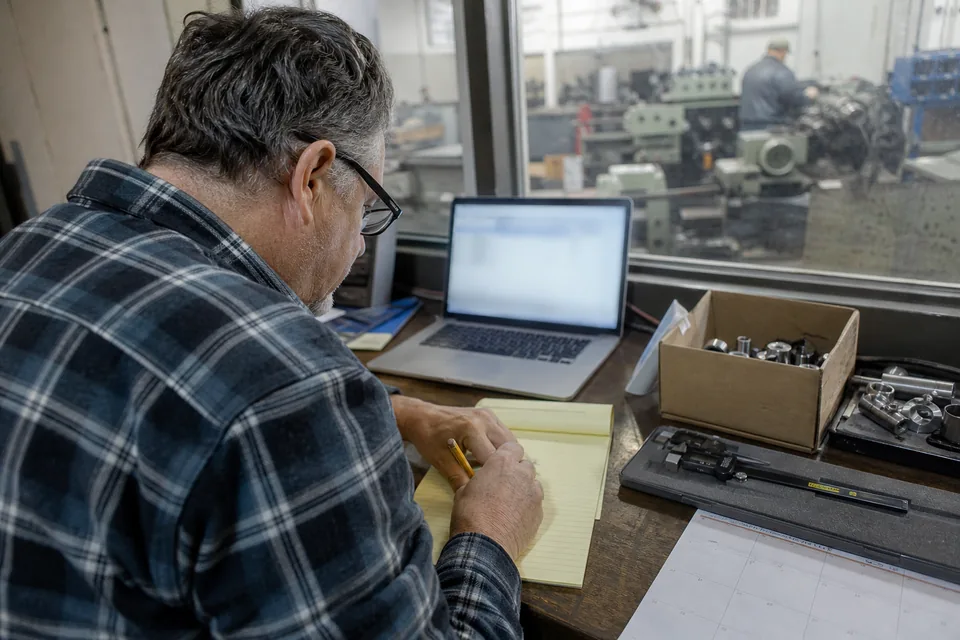Machine shop owner writing on a notepad beside a laptop in his front office, lathes visible through the interior window