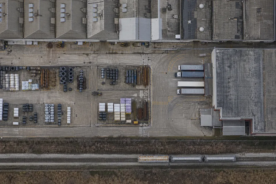 Drone view straight down on a manufacturing plant with sawtooth roofs, a rail siding, stacked steel coils and pallets in the yard
