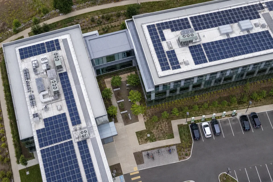 Drone view straight down on a glass office campus roof with rows of solar panels, a courtyard with benches and a visitor parking lot