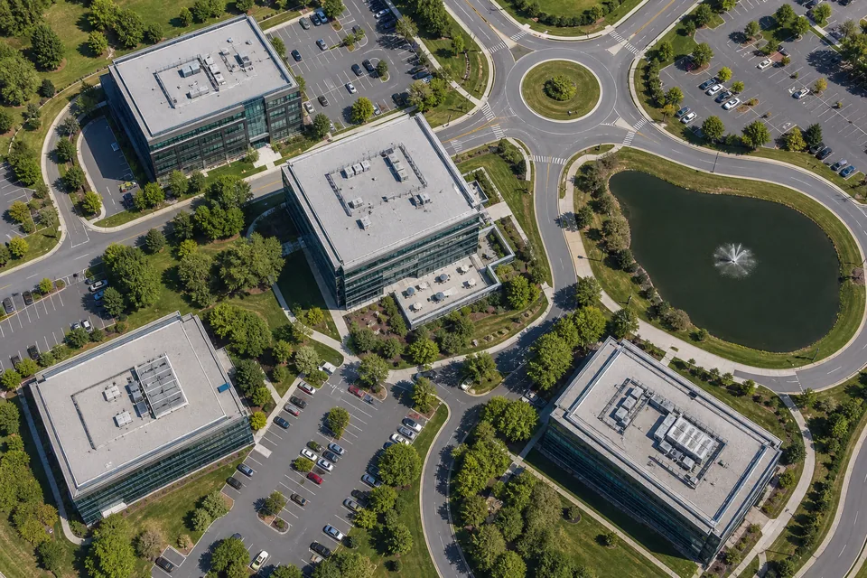 Drone view straight down on a suburban corporate office district with low-rise glass office buildings, landscaped courtyards, tree-lined walkways, a half-full employee parking lot and a retention pond
