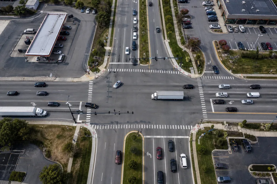 Drone view straight down on a busy US road junction with cars and trucks moving in marked lanes, turn lanes, crosswalk stripes and grass medians