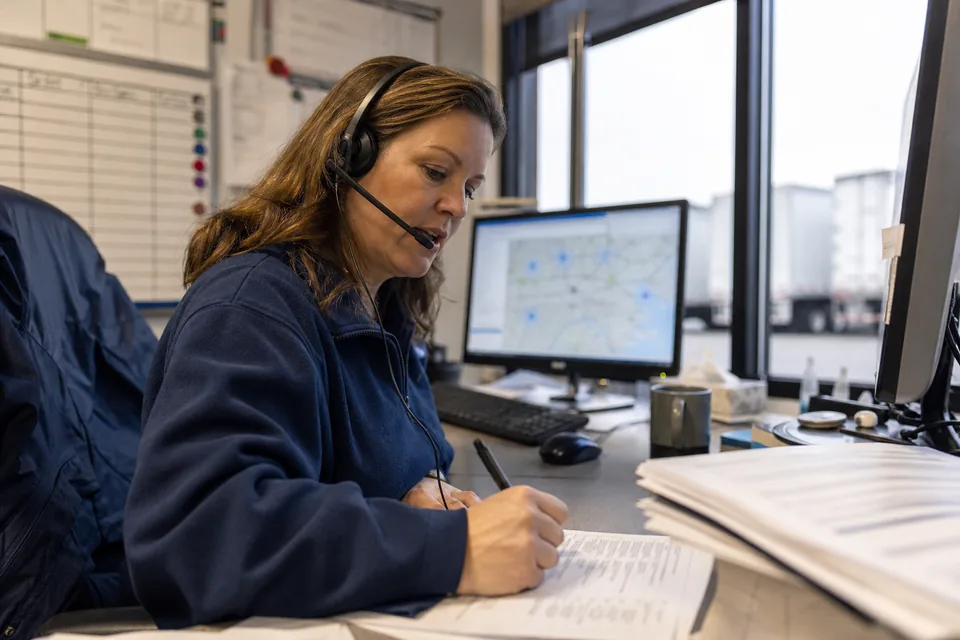 A regional sales rep at her desk in a logistics company office, headset on, marking a printed account list with a pen, a dispatch board and a window to the truck yard behind her