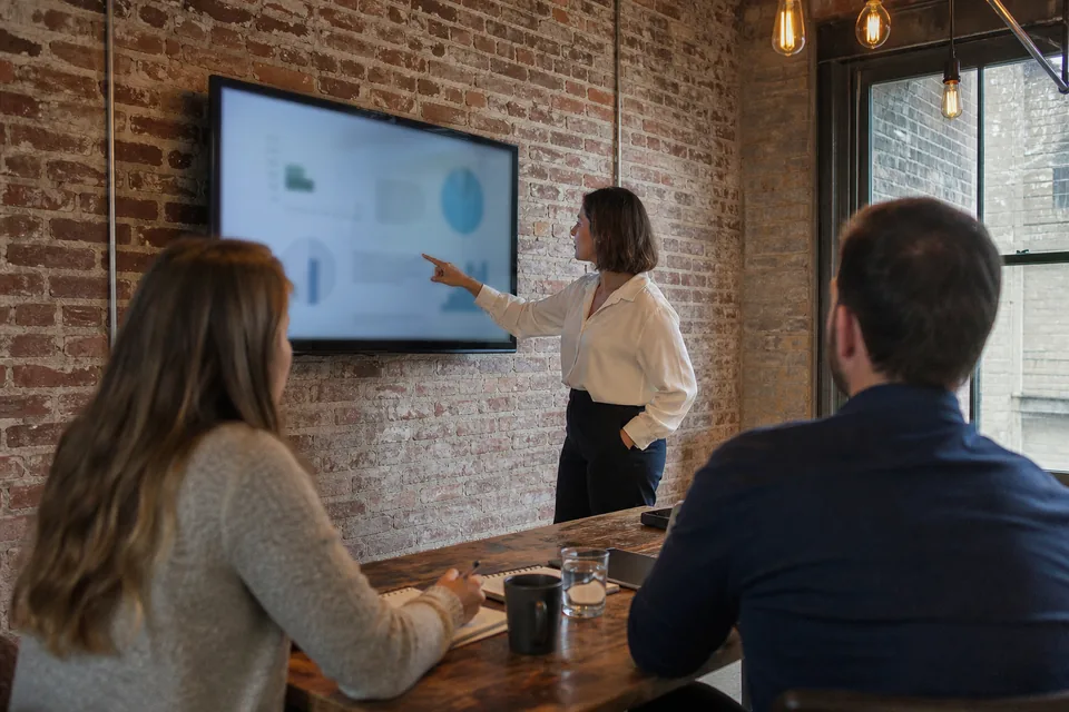 Agency strategist presenting a blurred report on a wall screen to clients seated at a wooden table in a brick loft