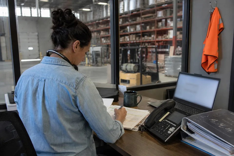 Sales rep at a warehouse office standing desk marking a printed call list with a pen, a headset on the desk and a forklift visible through the window