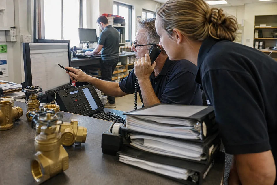 Inside sales rep at an industrial supply distributor on the phone, pointing at a list on his monitor while a colleague leans in, valve samples and catalog binders on the counter