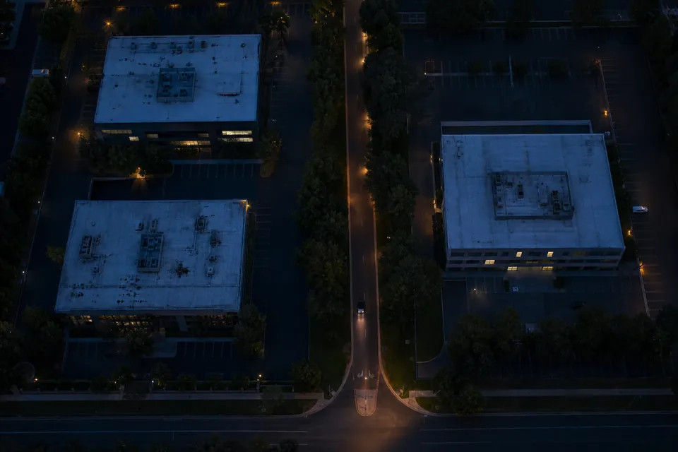 Drone view straight down on a quiet street of office buildings at dusk with lit windows, empty parking spaces and warm street lamps